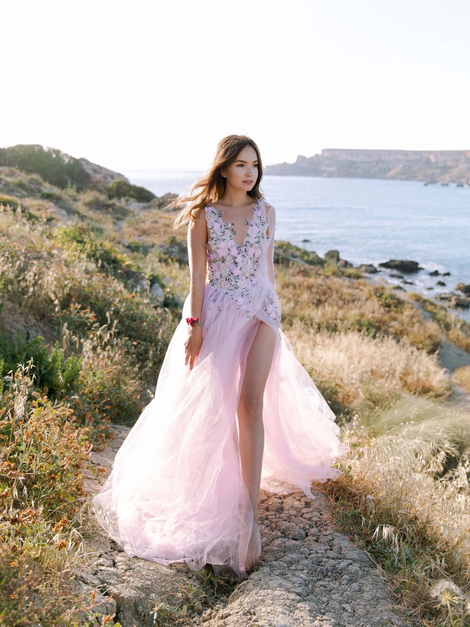 Bride in a blush tulle gown walking a stony path through dry grass above the sea, Maltese coast Bride in a blush tulle gown walking a stony path through dry grass above the sea, Maltese coast