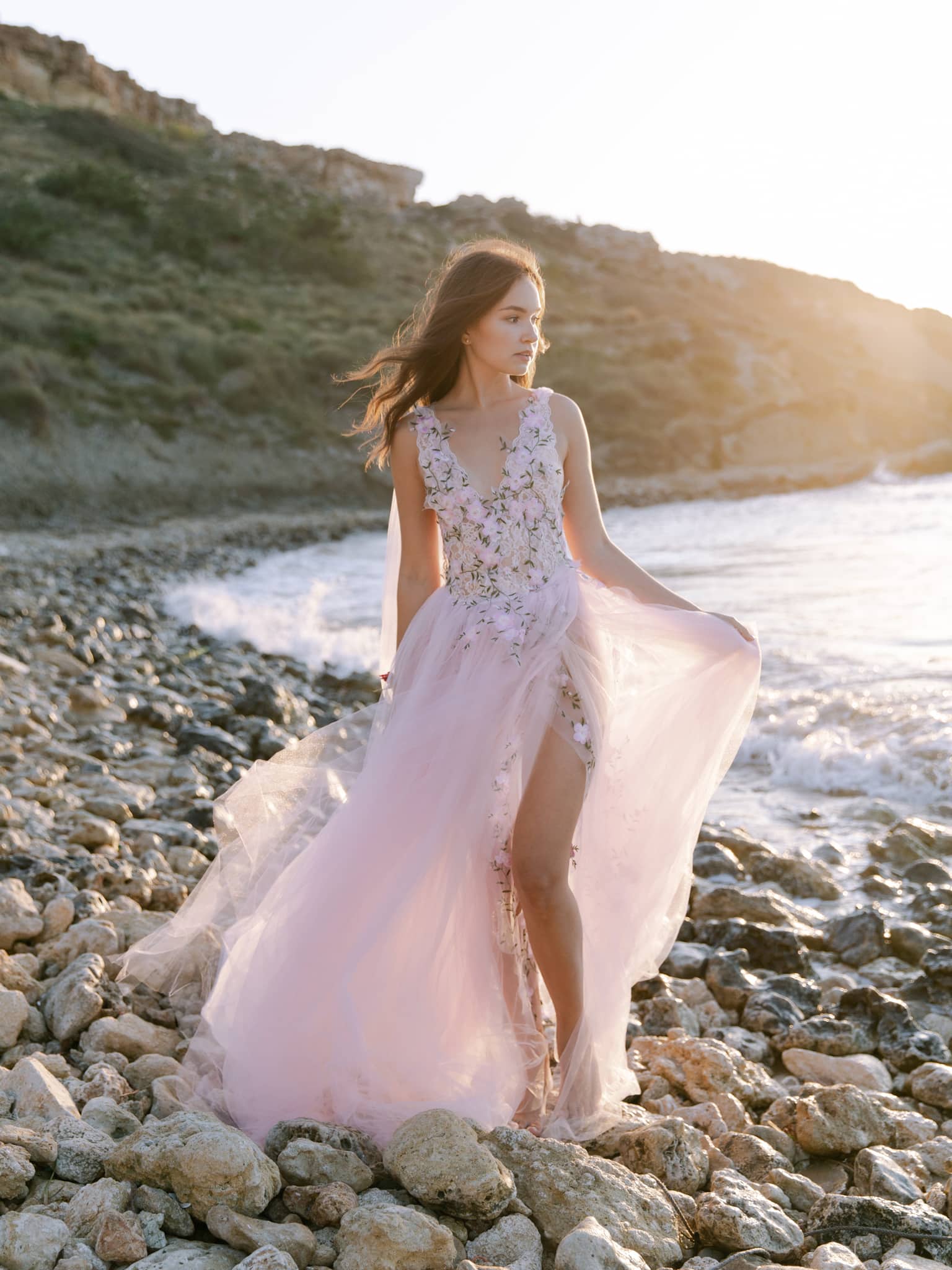Bride on a stony beach holding out her tulle skirt as waves break behind her, backlit by the low sun, Malta Bride on a stony beach holding out her tulle skirt as waves break behind her, backlit by the low sun, Malta