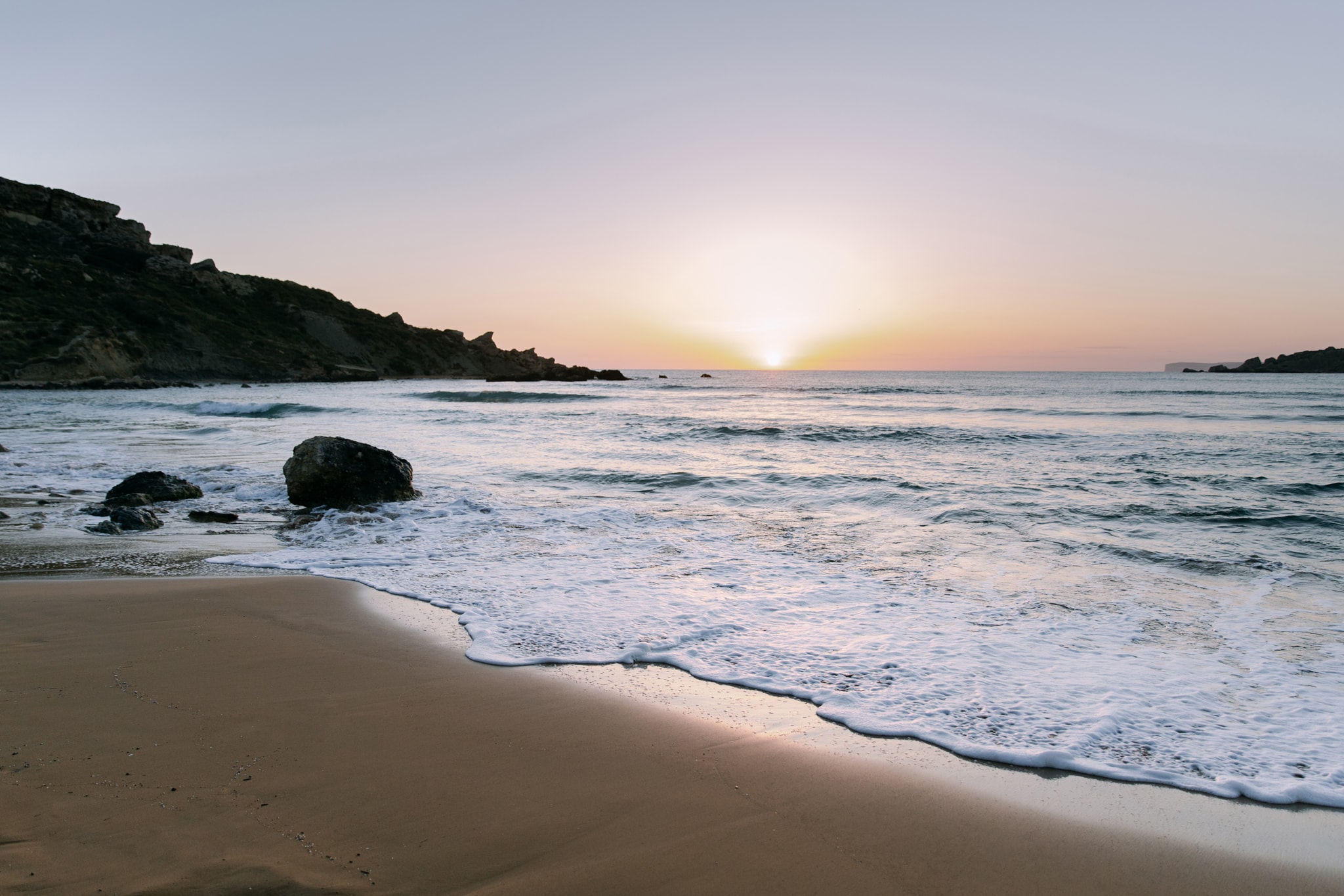 The sun setting into the sea beside a headland, seen from a sandy beach in Malta The sun setting into the sea beside a headland, seen from a sandy beach in Malta