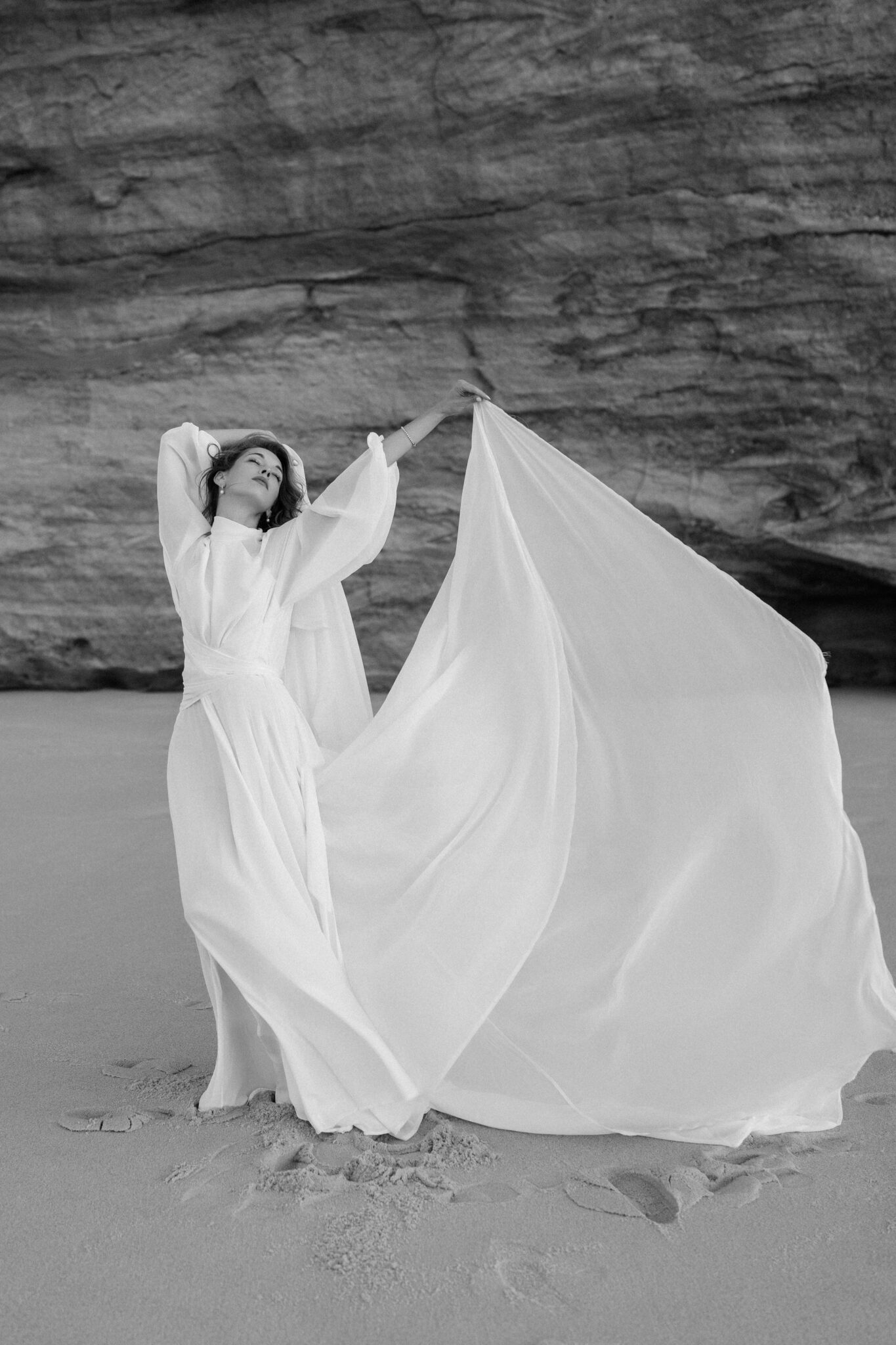 Bride holding her long skirt up in the wind in front of the layered rock wall at Praia del Rey, black and white Bride holding her long skirt up in the wind in front of the layered rock wall at Praia del Rey, black and white
