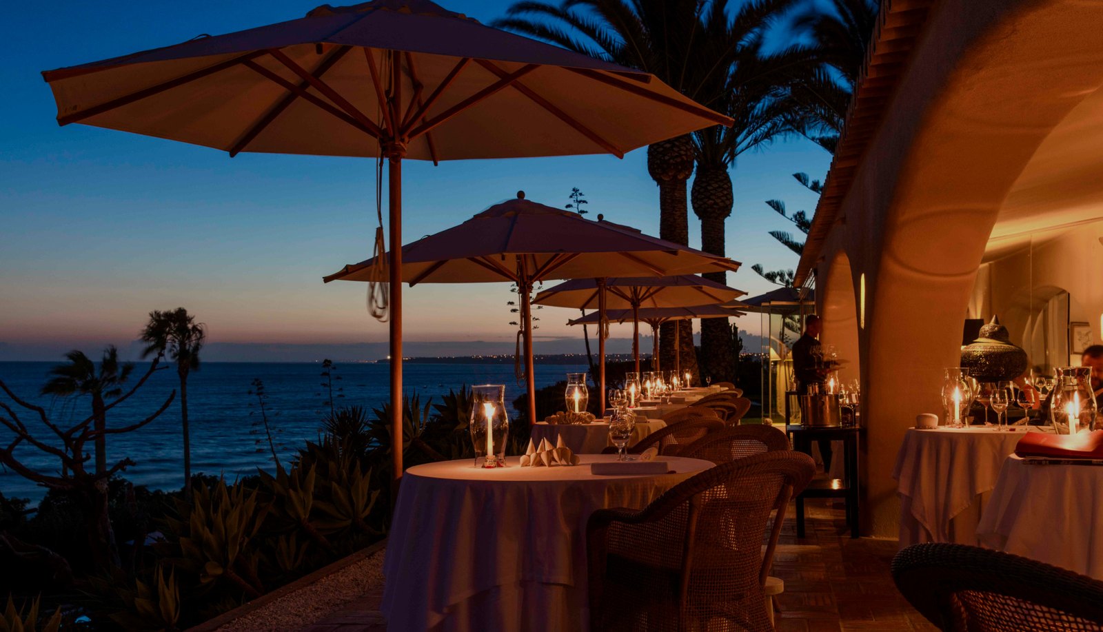 Candlelit tables under parasols on the terrace above the sea at dusk, Vila Joya, Praia da Galé Candlelit tables under parasols on the terrace above the sea at dusk, Vila Joya, Praia da Galé