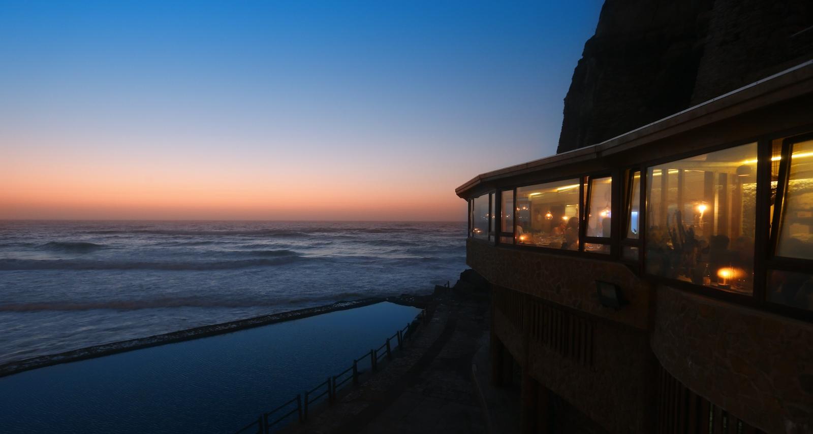 The glass-walled restaurant lit at dusk above the ocean pool, Azenhas do Mar, Sintra coast