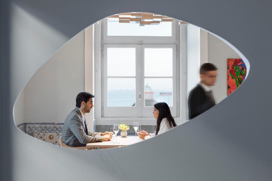 A couple at a table seen through an oval opening in a white wall, a window onto the river behind them, at Epur, Lisbon