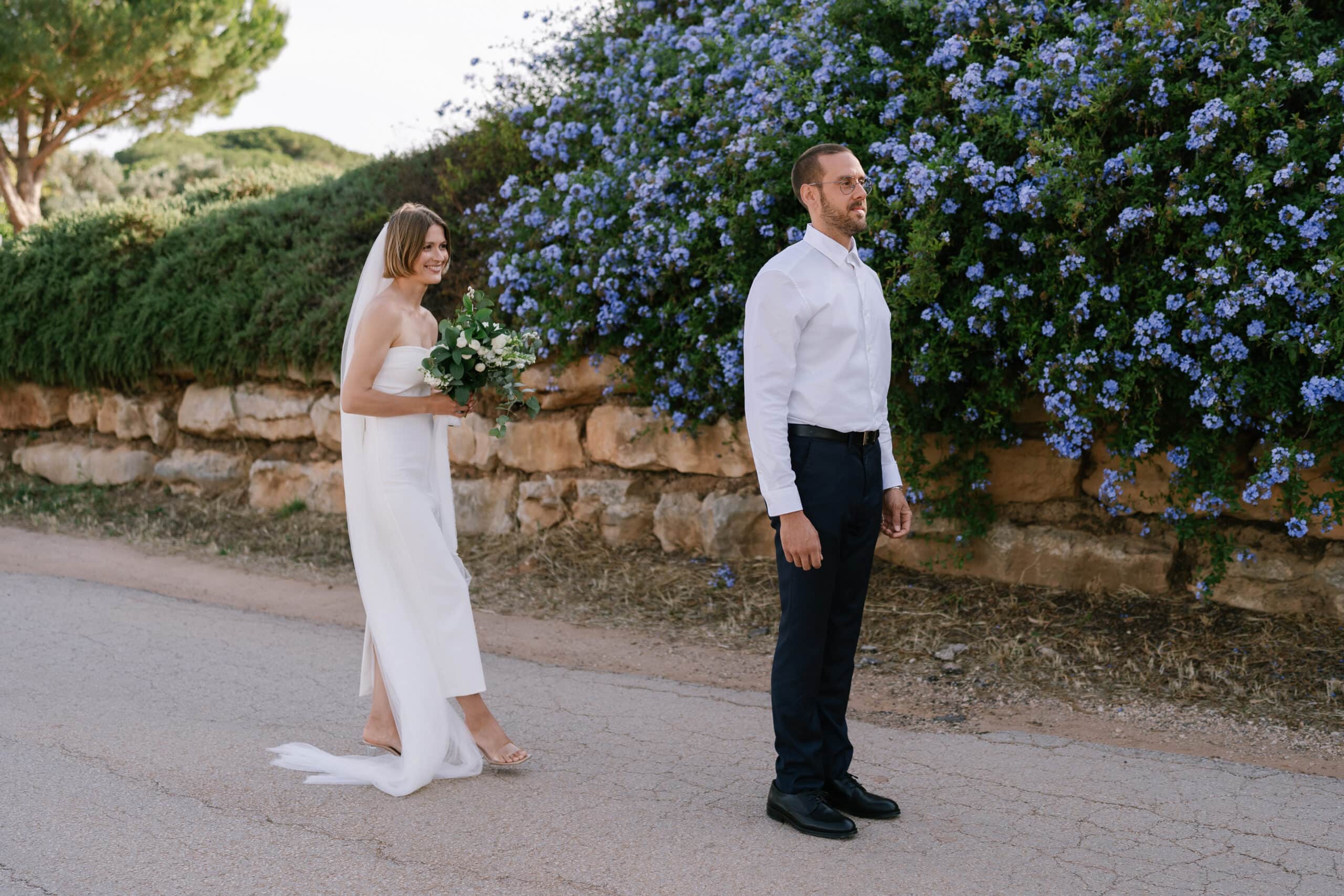 Bride approaching groom for a First Look at their Algarve elopement, with a backdrop of blooming blue flowers.