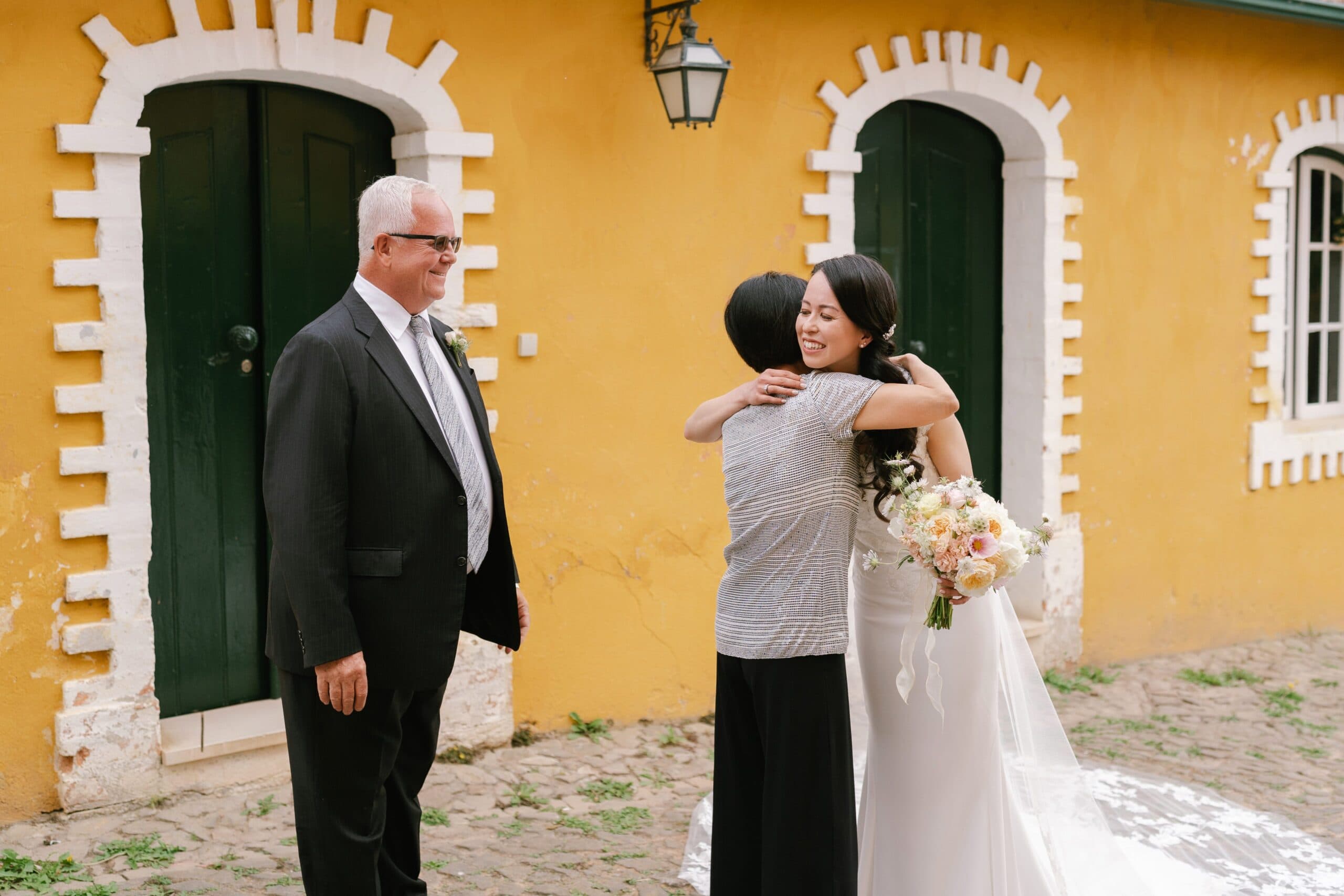 Bride having a first look with her parents, sharing a joyful embrace against a vibrant yellow background.