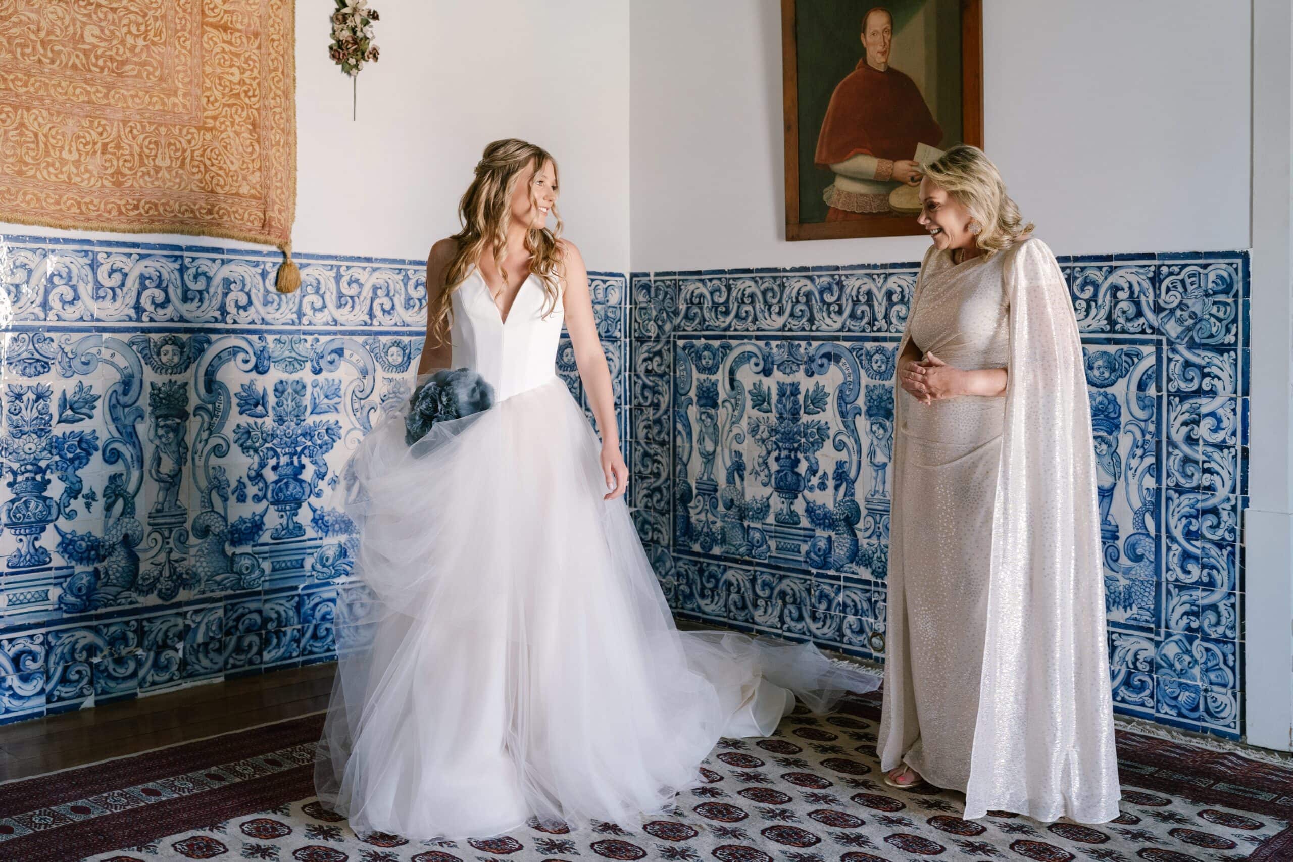 Bride in a flowing white gown sharing a heartfelt First Look with her mother in a glittering gold dress, in the traditional azulejo-tiled room of Palacio de Correio-Mor in Portugal