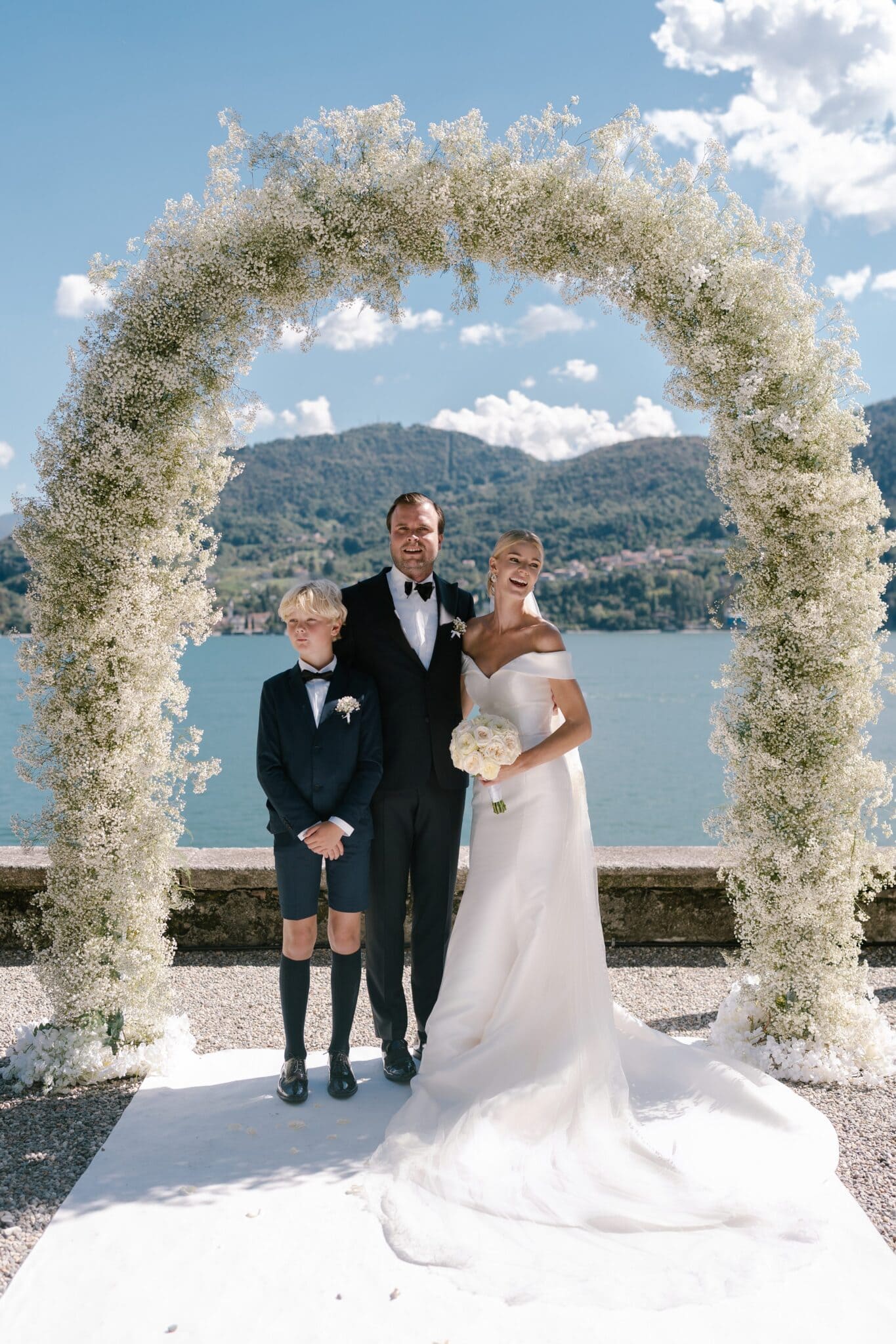 Bride and groom with a young boy standing under a floral arch overlooking Lake Como at Villa Carlotta.