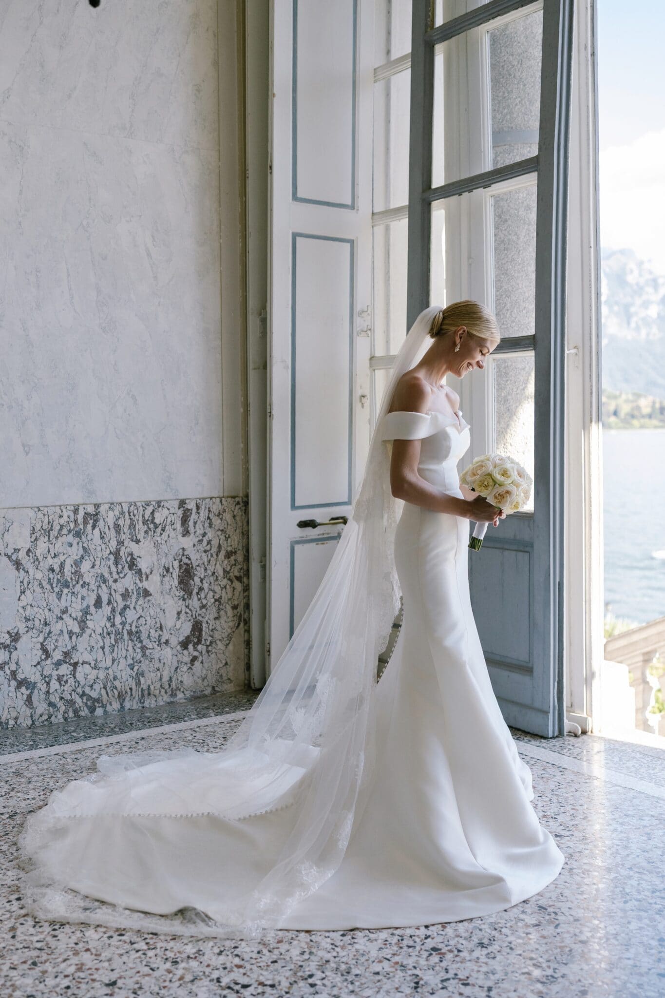 Bride in a fitted off-the-shoulder gown holding a white rose bouquet by a window at Villa Carlotta, Lake Como.