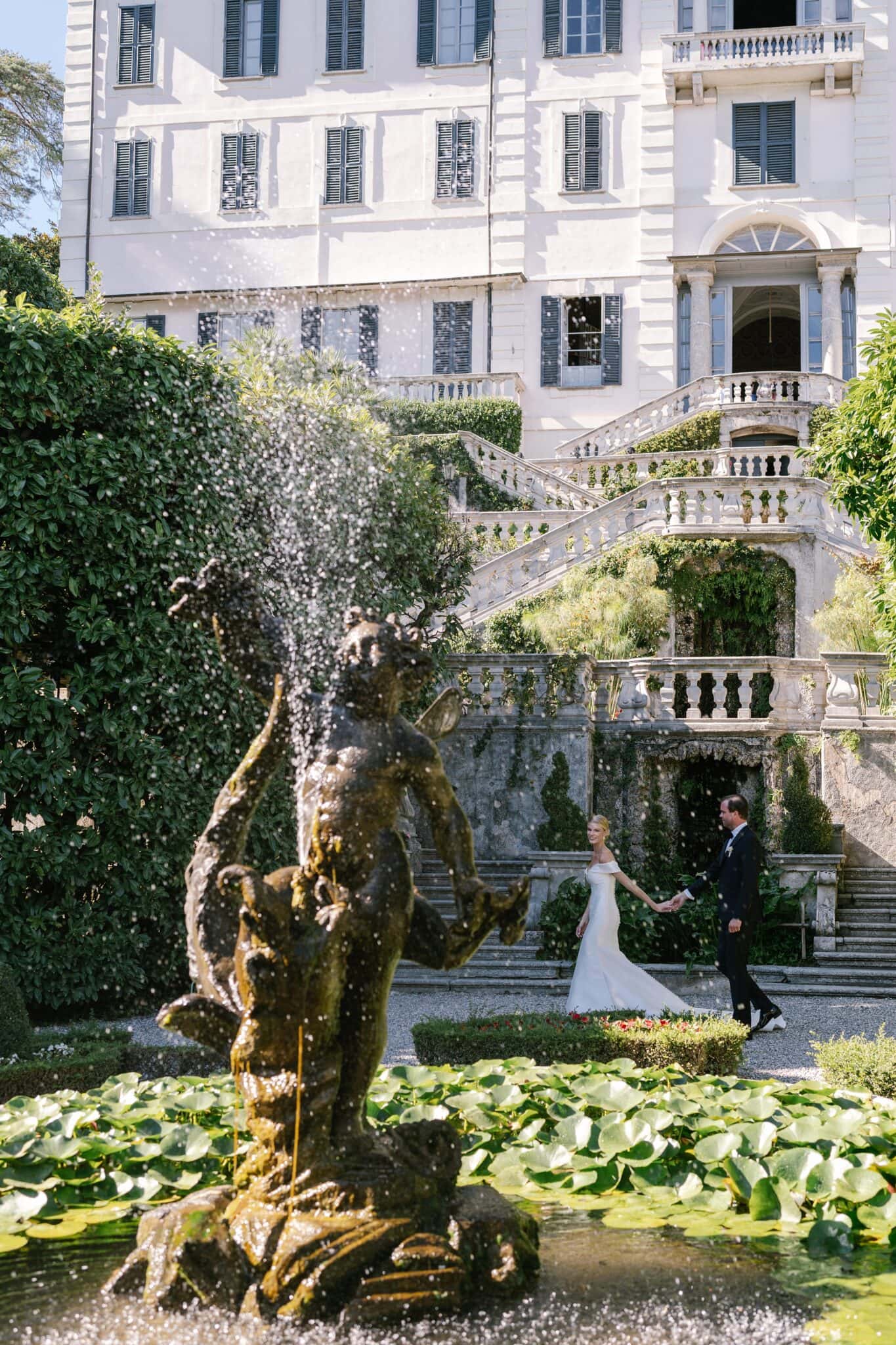 Bride and groom walking hand in hand in Villa Carlotta gardens, framed by the fountain and greenery overlooking Lake Como.