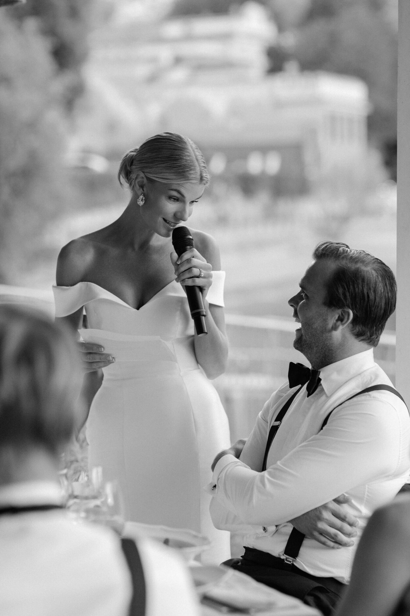Bride giving a wedding speech with a microphone as the groom smiles during the reception at Lido di Lenno, Lake Como.