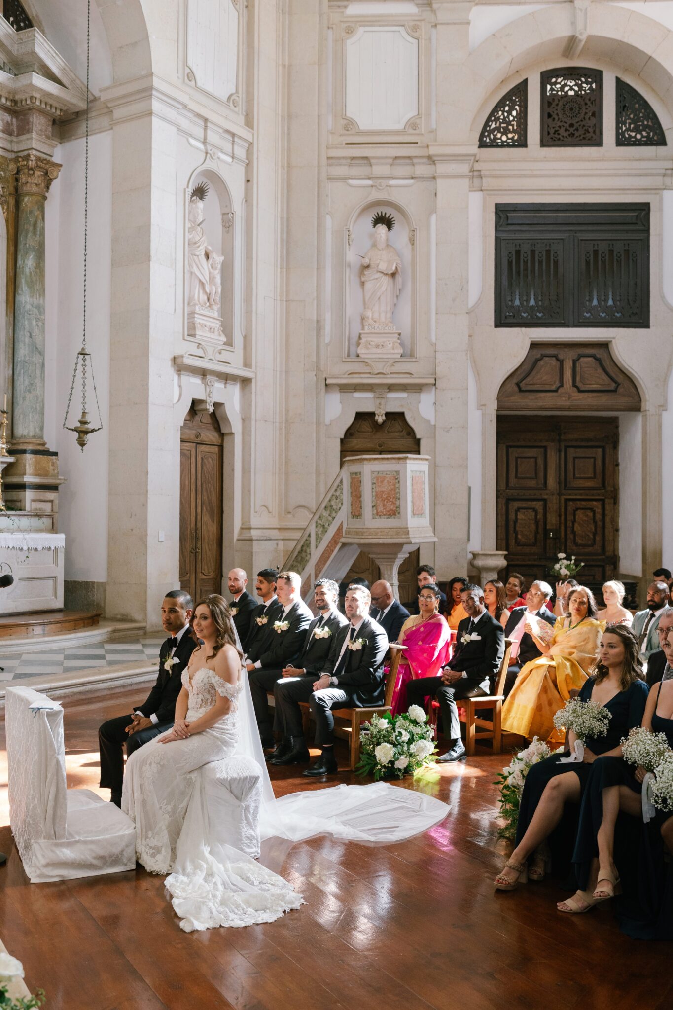Bride and groom seated at altar during church ceremony surrounded by wedding guests in Óbidos Portugal