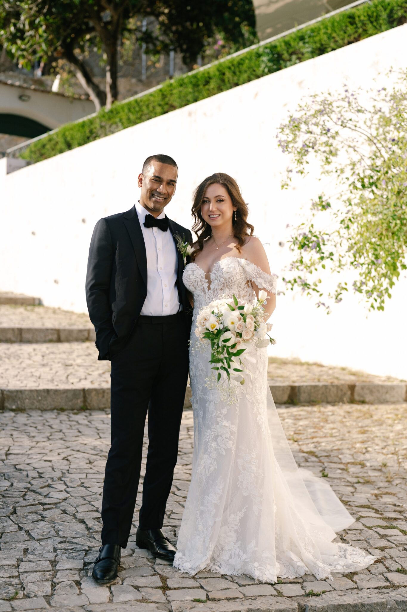 Bride and groom standing together on cobblestone steps with whitewashed walls and greenery in Óbidos, Portugal