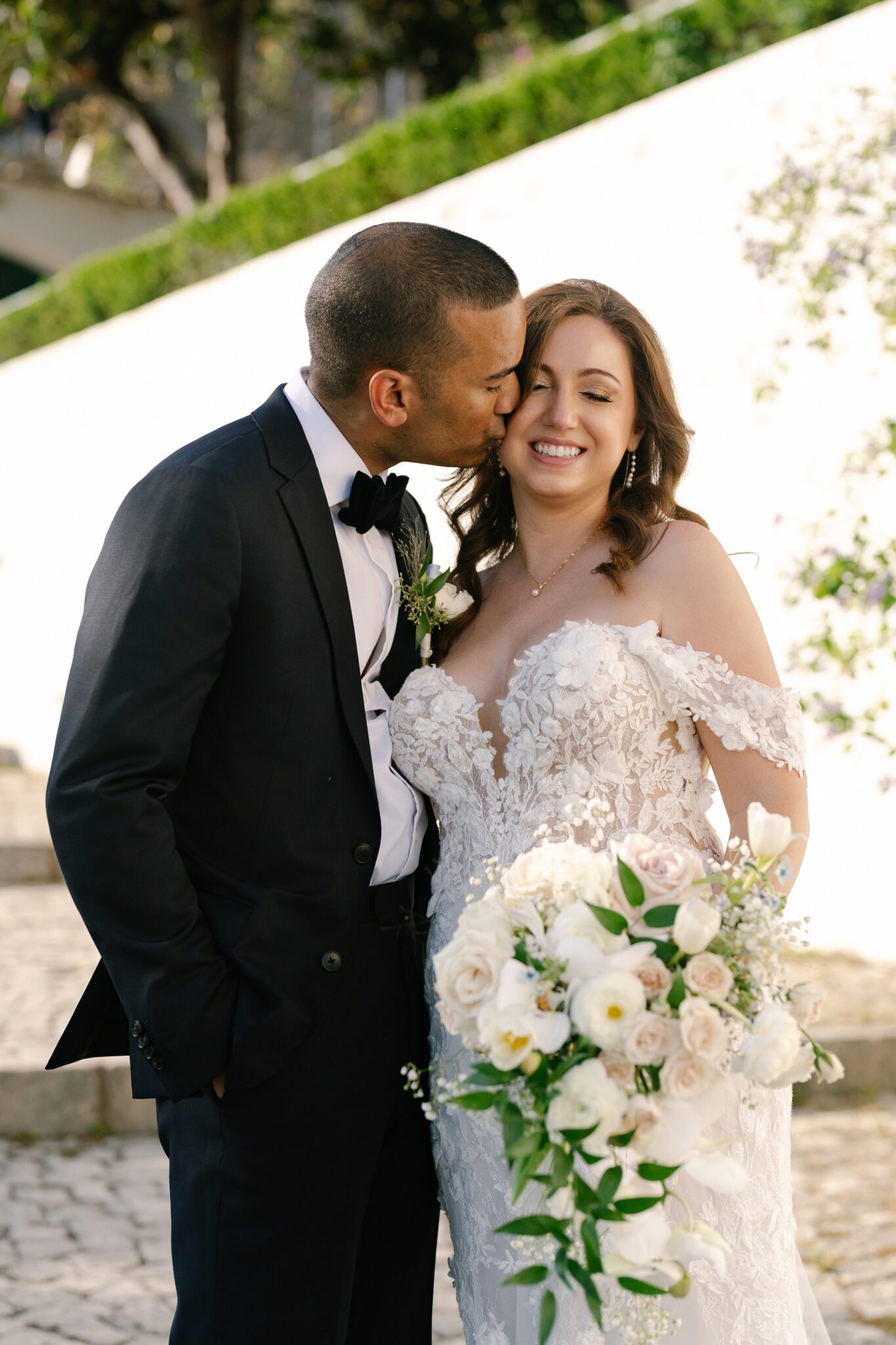 Groom kissing his smiling bride in Óbidos, Portugal, as she holds an elegant bouquet