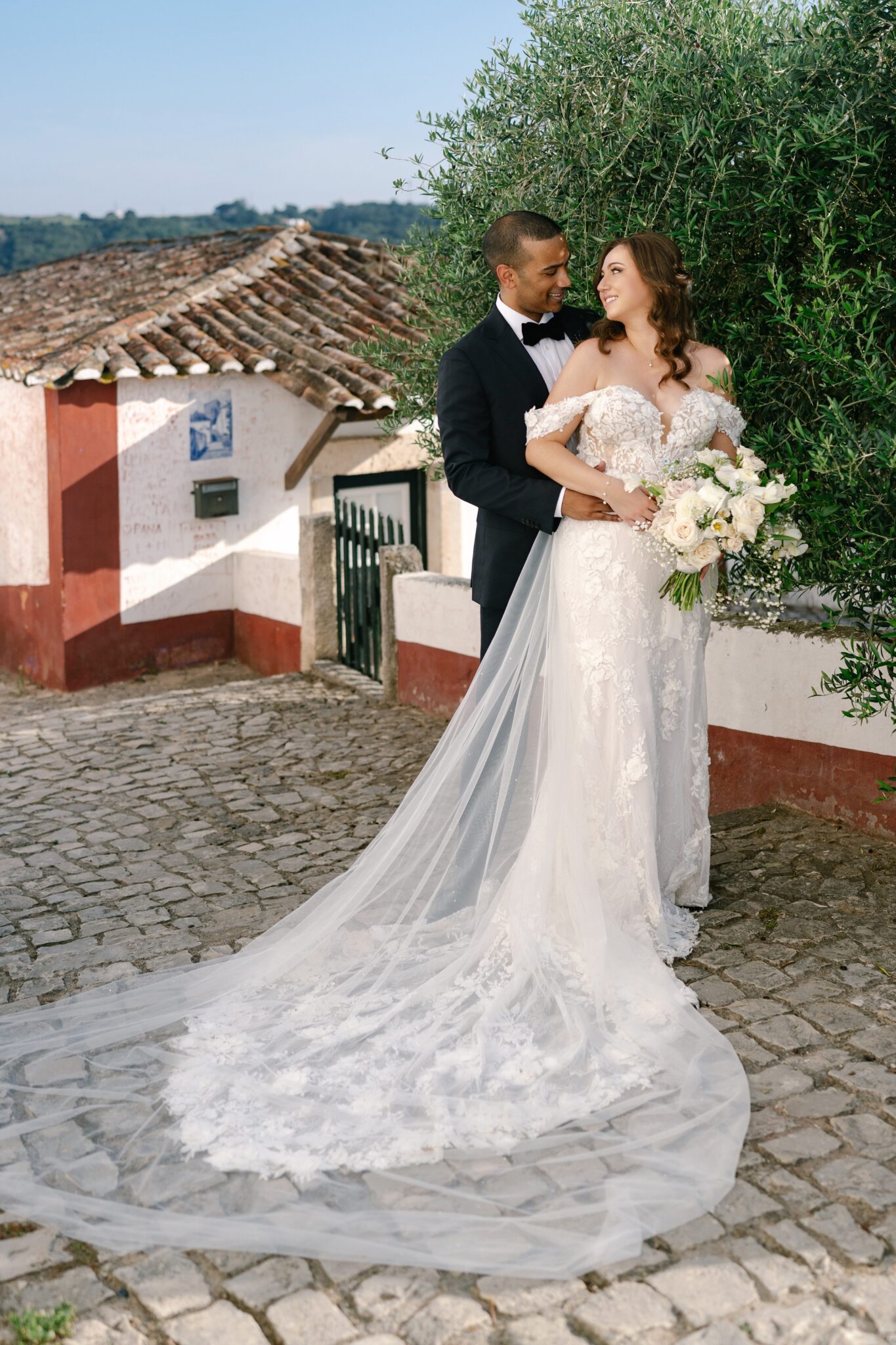 Bride and groom embracing by olive tree and rustic wall in Óbidos Portugal