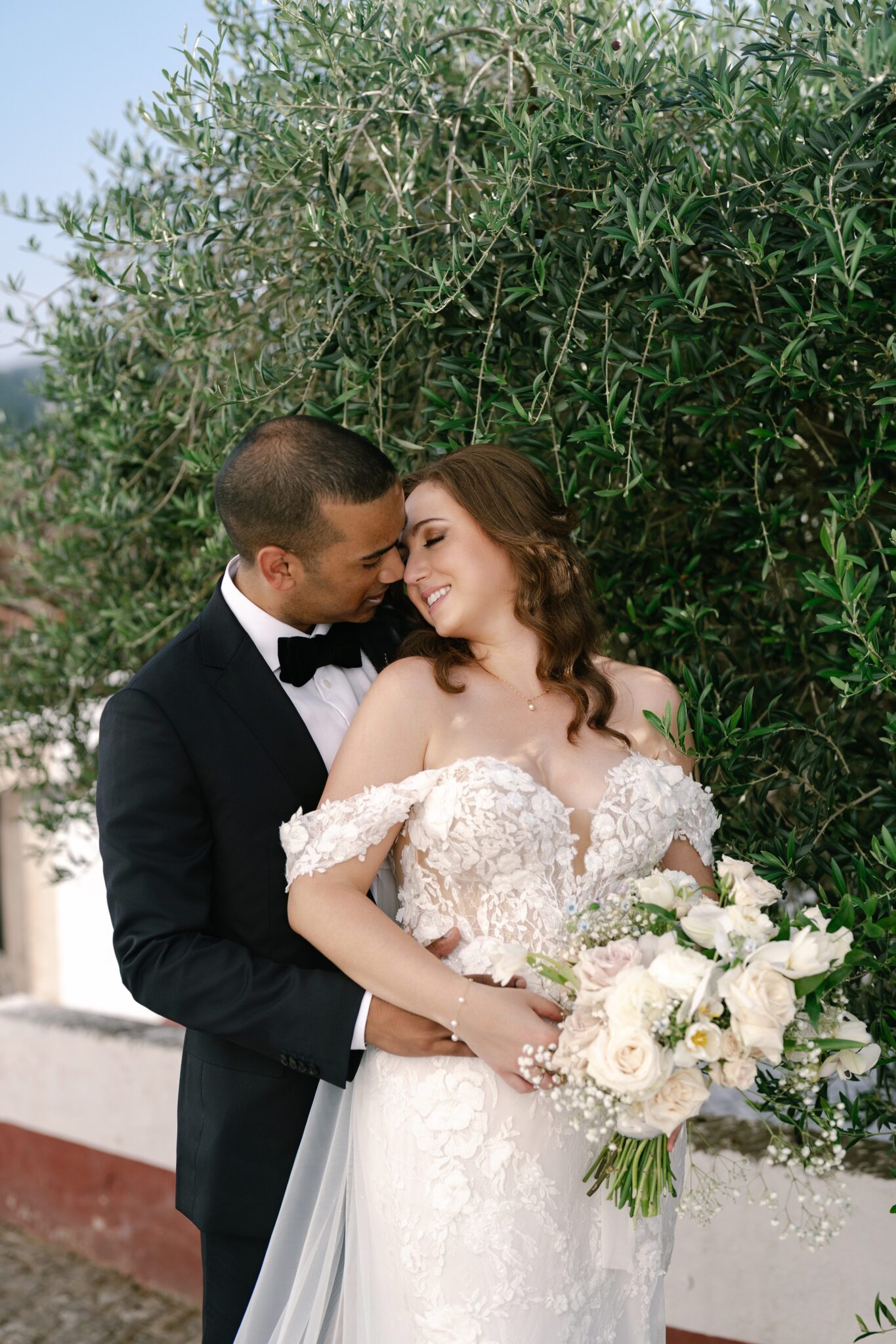 Groom embracing bride with bouquet under green tree in Óbidos Portugal