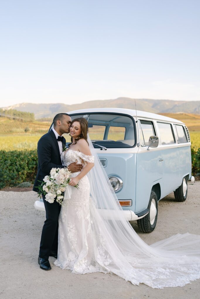 Groom kissing bride’s forehead beside a vintage blue VW van at Quinta do Gradil vineyard, Portugal