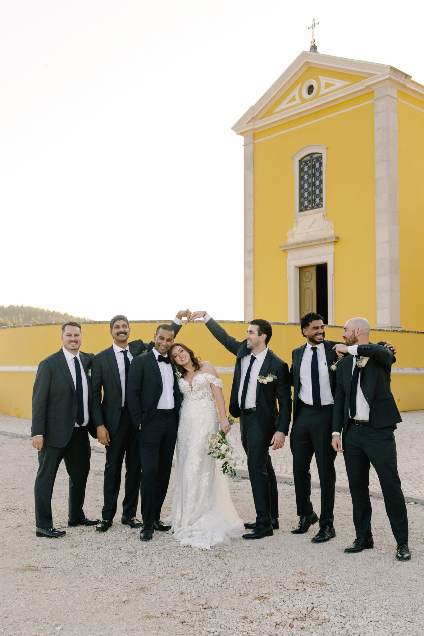 Bride and groom laughing with groomsmen in front of yellow chapel at Portugal wedding